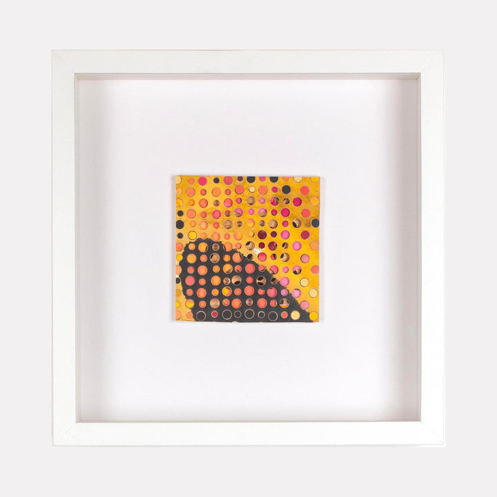 The artwork 'Chisholn' by Margaret Griffith features a vibrant square composition with polka dot patterns on cut paper. A golden yellow background transitions to deep charcoal, decorated with multicolored circular elements in pink, orange, and black. The piece is framed in crisp white, creating a striking contrast. Acrylic on cut paper, by Margaret Griffith.