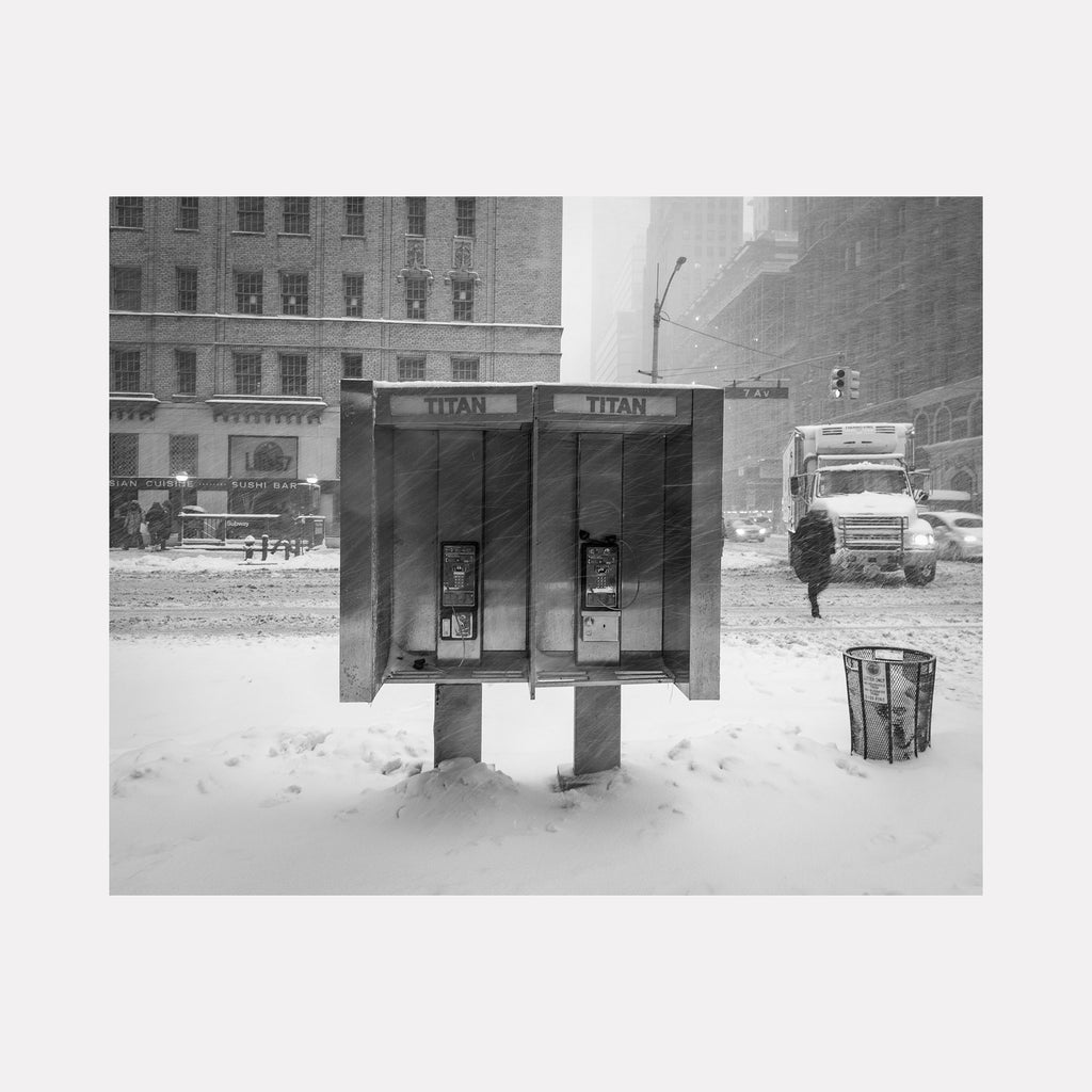 The artwork 'Cold Call' by Dennis Maida captures a black and white urban photograph of two phone booths in a snowy cityscape. The vintage Titan phone booths stand isolated against a backdrop of classic brick buildings, with gentle snowfall creating a moody winter atmosphere. A trash bin and distant truck complete the nostalgic street scene.