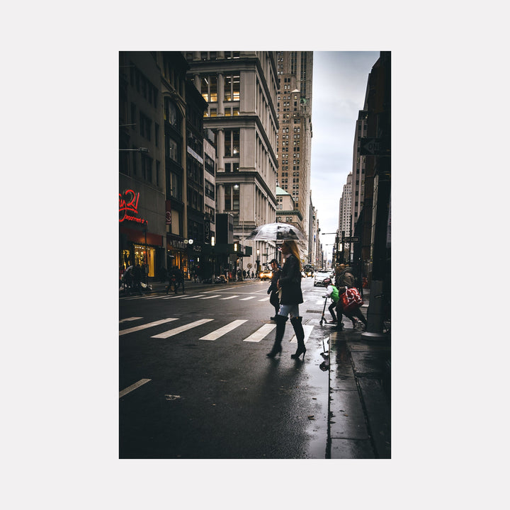 The artwork 'Daily Commute' by Dennis Maida captures an atmospheric urban street scene in moody tones. A silhouetted figure crosses a rain-slicked crosswalk against towering city buildings. Golden shop lights glow warmly through the dim cityscape, while vintage architecture and fire escapes create dramatic vertical lines. The photograph's composition draws the eye down the shadowy canyon of skyscrapers and storefronts.