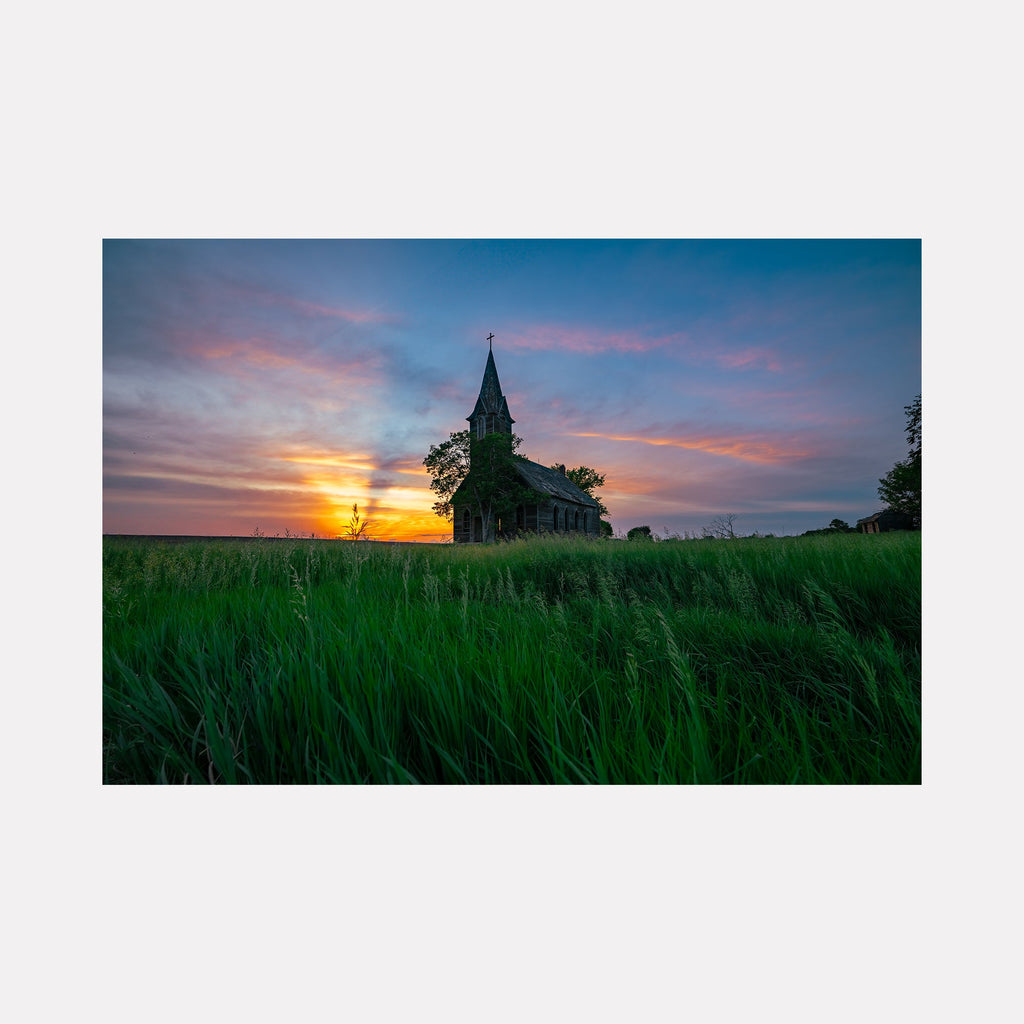 The artwork 'Evening Prayer' by Dennis Maida captures a Gothic church silhouetted against a dramatic sunset. The composition features vibrant green prairie grass in the foreground, a weathered wooden church with a distinctive spire, and a mesmerizing sky painted in hues of orange, pink, and deep blue. The photographic artwork beautifully portrays the peaceful solitude of rural architecture at dusk.