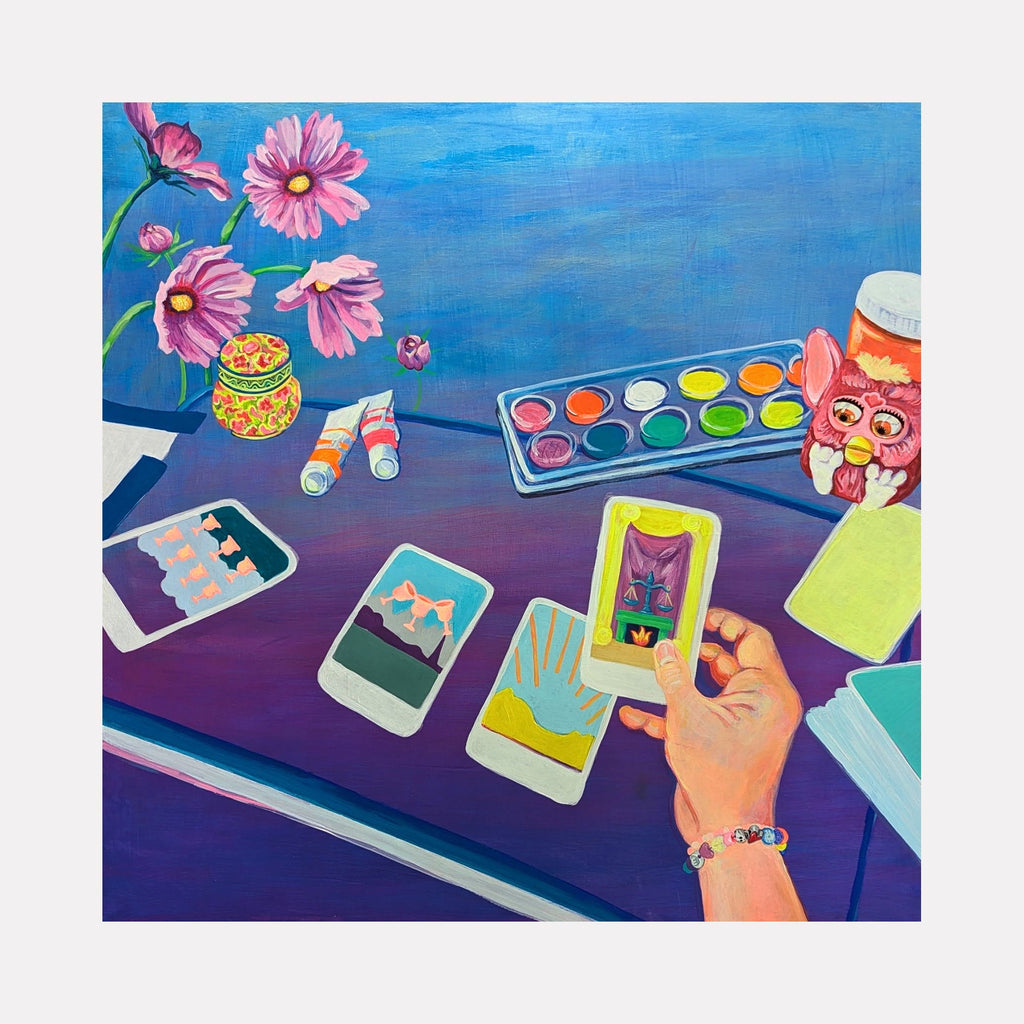 The artwork 'Justice in Her Hands' by Mary Younkin features a vibrant workspace with a watercolor palette, pink cosmos flowers in bloom, and instant photos laid out on a purple surface. A hand holds a phone displaying artistic content, while decorative jars and art supplies create a creative atmosphere. Painted in acrylic on wood panel, the piece combines bright blues, pinks, and purple hues. By Mary Younkin.