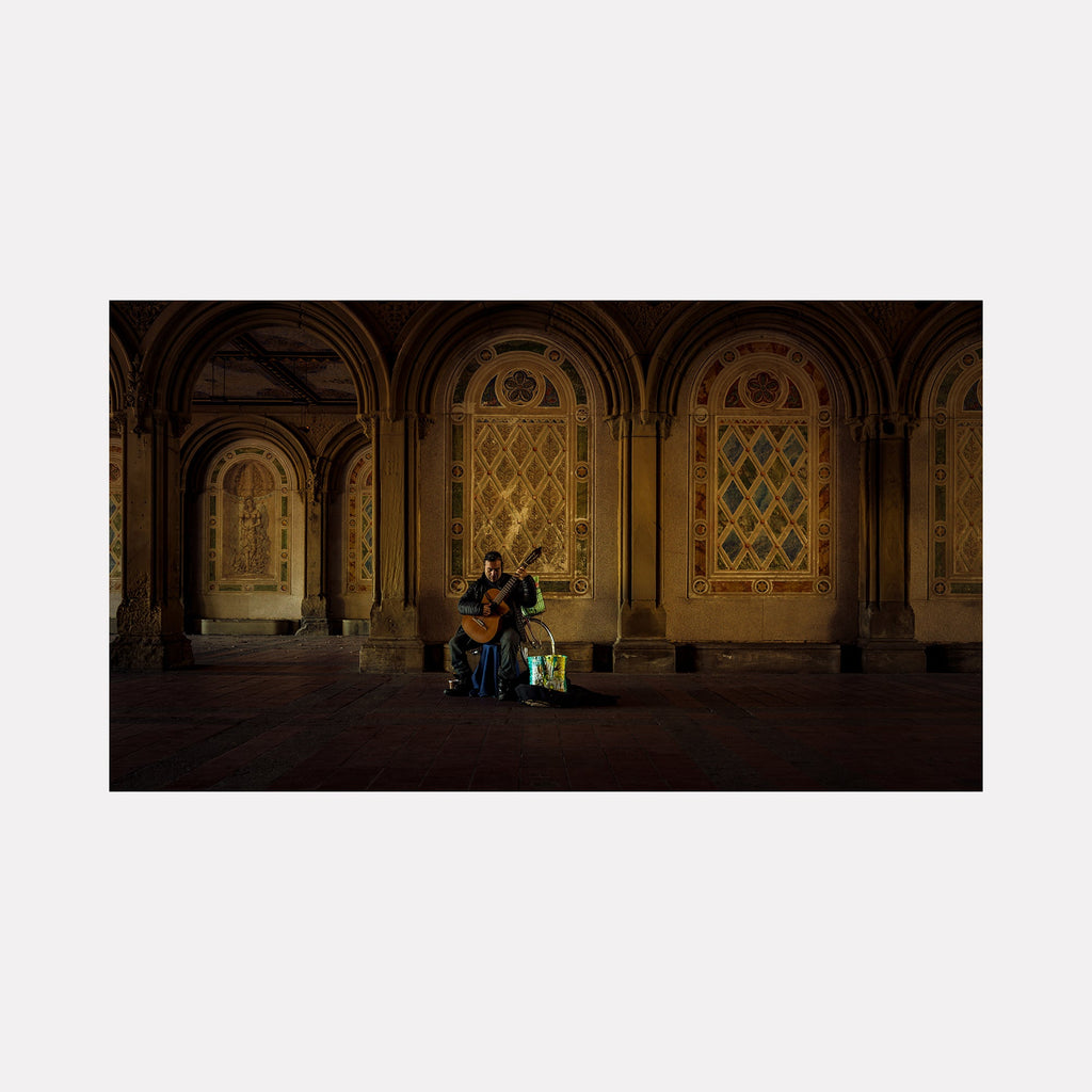 The artwork 'Maestro' by Dennis Maida captures a musician performing in an ornate architectural setting, featuring dramatic gothic arches and intricate latticed windows illuminated in warm golden light. The composition showcases historic stone archways with decorative details against a dark atmospheric background, creating a moody nighttime scene in Central Park's Bethesda Terrace. By Dennis Maida.