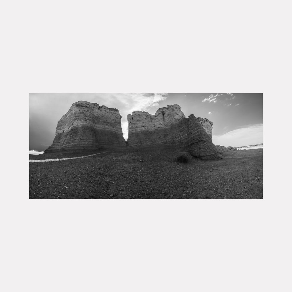 The artwork 'Monuments of Time' by Dennis Maida captures a dramatic black and white panoramic photograph of weathered limestone formations against a stark desert landscape. Towering rock monuments with layered striations cast deep shadows on the barren ground, while wispy clouds drift across the contrasting sky, creating a timeless geological portrait. Photograph by Dennis Maida.