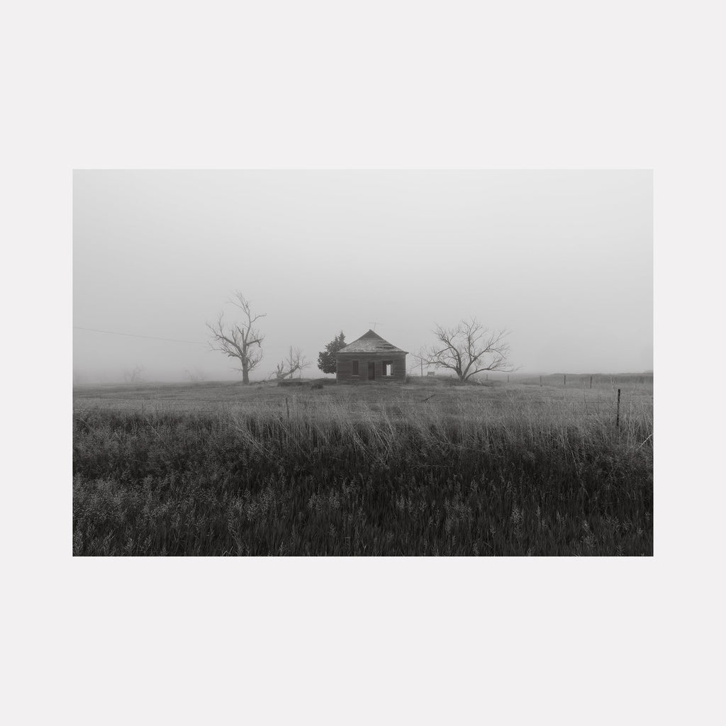 The artwork 'Nevermore' by Dennis Maida captures a haunting black and white photograph of an abandoned farmhouse standing solitary in a misty prairie landscape. Bare trees flank the weathered structure, while tall prairie grass dominates the foreground, creating a moody atmospheric composition suggesting isolation and the passage of time. Photograph by Dennis Maida.