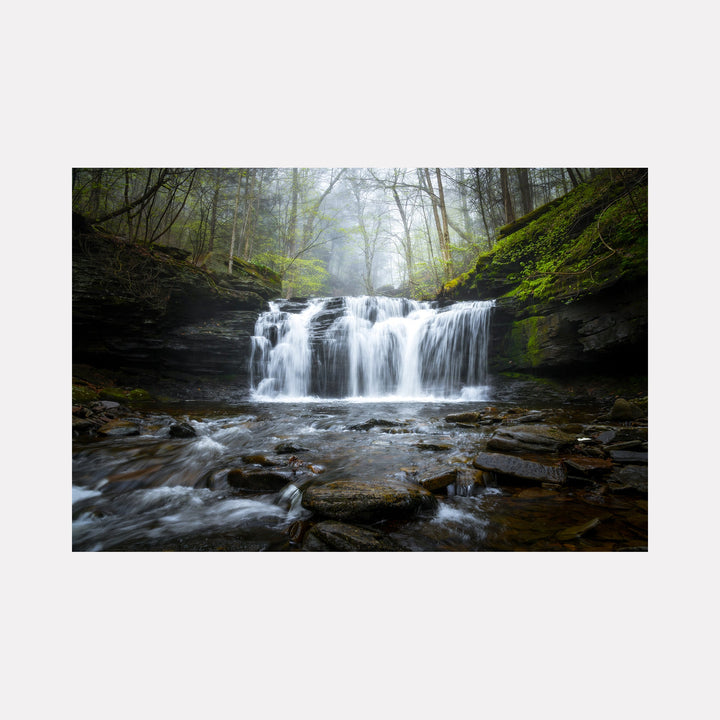 The artwork 'The Music of Water' by Dennis Maida captures a serene waterfall cascading over dark rocky ledges in a misty forest setting. Silky white water flows over multiple tiers of slate-gray rock, surrounded by moss-covered stones and bare winter trees in soft morning fog. The photograph masterfully captures the movement and ethereal quality of flowing water against the rugged natural landscape.