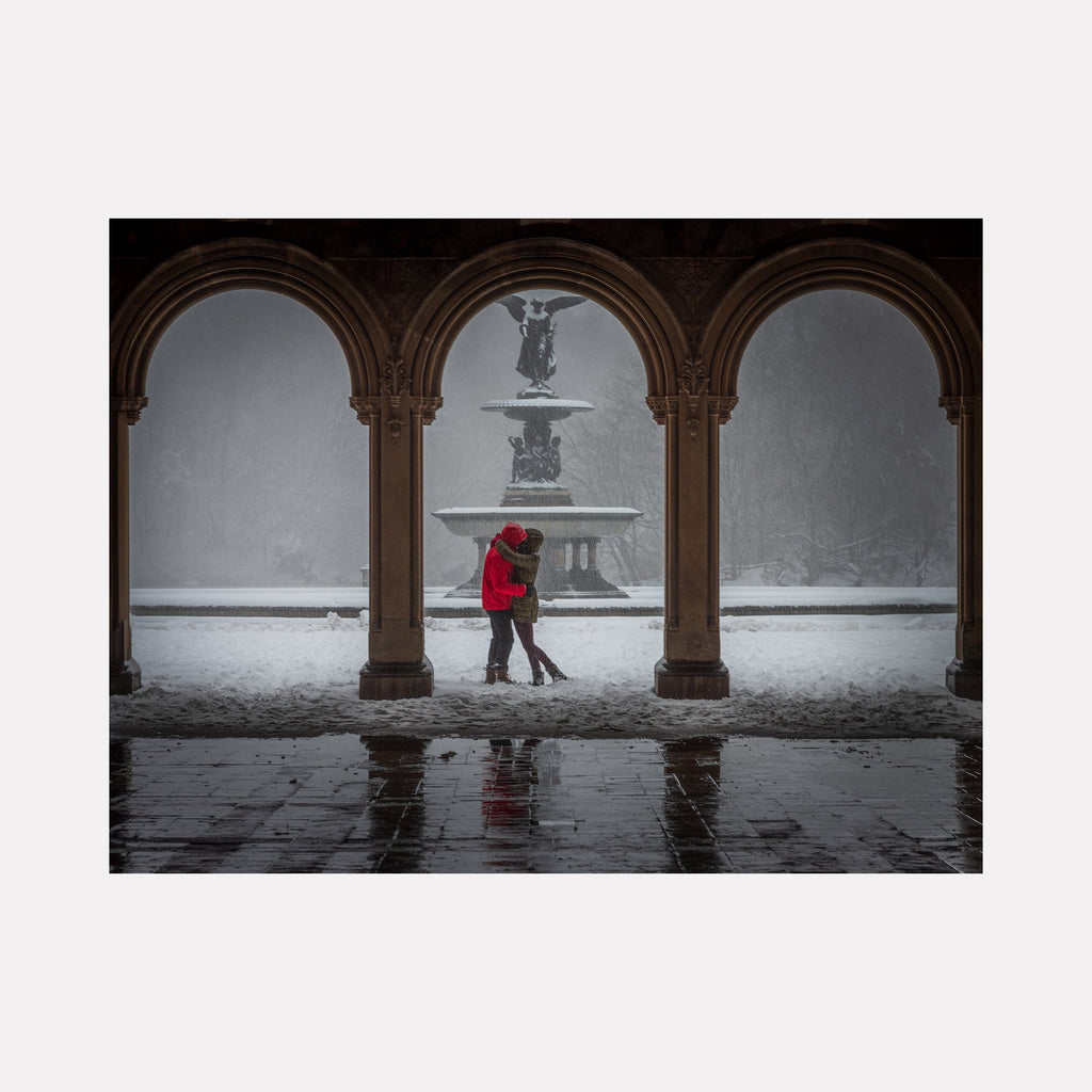 Two Souls Beneath the Winter Arch by Dennis Maida captures a romantic winter scene through ornate stone archways in Central Park. A figure in a vibrant red coat stands near the snow-covered Bethesda Fountain. The gothic architecture frames the misty winter landscape, with reflected arches on wet pavement creating dramatic symmetry. Photograph, 36x27 inches.