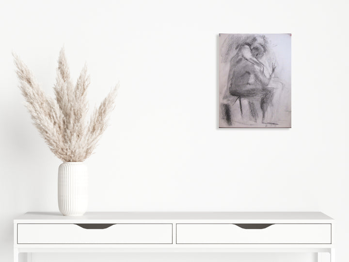 The artwork 'Claudia and Valentine' by Peter Colquhoun features expressive charcoal strokes on paper depicting intimate forms, mounted on a white wall above a modern console table with dried pampas grass in a ribbed white vase. The 15x12 inch drawing showcases gestural mark-making and tonal variations characteristic of charcoal medium.