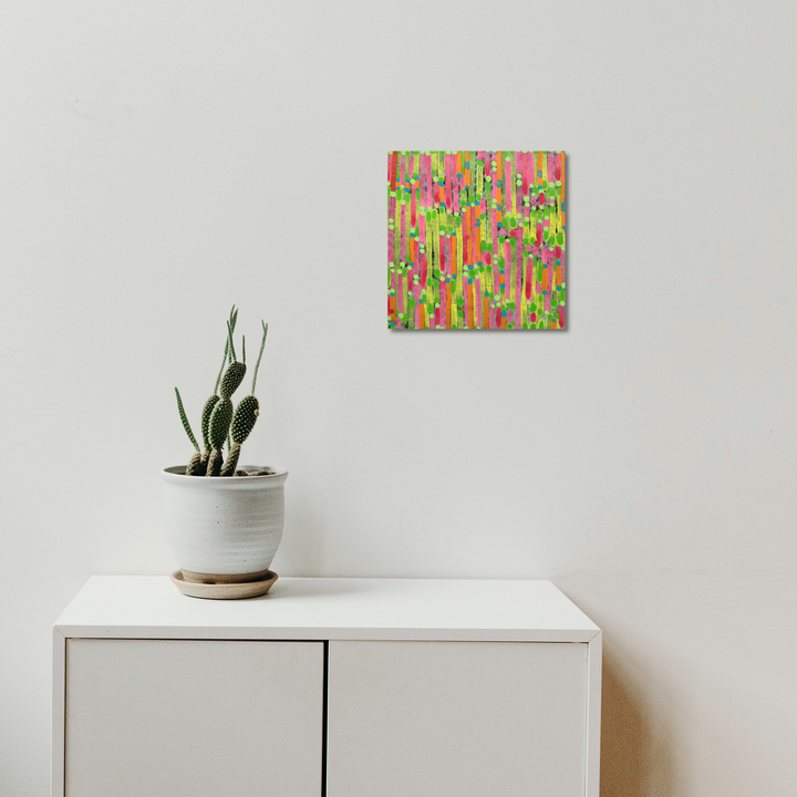 The artwork 'Composition No. 226' by Raul Ortiz features vibrant vertical brushstrokes in neon green, hot pink, and orange creating an abstract pattern on linen canvas. The 12x12 inch acrylic painting displays energetic movement through parallel lines, reminiscent of a tropical garden in full bloom, mounted above a white cabinet with a potted cactus.