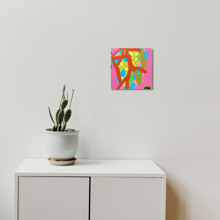 The artwork 'Composition No. 45' by Raul Ortiz features vibrant abstract shapes in pink, yellow, and green against a hot pink background. The 10x10 inch acrylic painting on cradled panel showcases organic flowing forms with geometric patterns, creating a dynamic modern composition. The artwork is displayed above a white cabinet with a potted cactus.