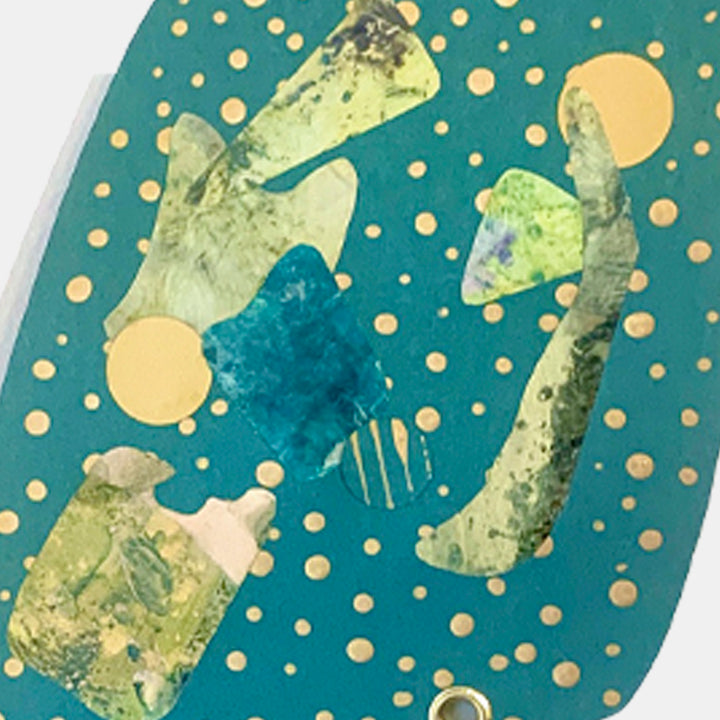 The artwork 'Dotter' by Sunny Chapman features abstract shapes in pale green and yellow floating on a deep teal background, adorned with golden polka dot pattern. Mixed media collage combines metallic paper and monotype prints, creating ethereal forms that seem to dance across the vertical composition. Metal grommets accent the contemporary piece's textural elements.