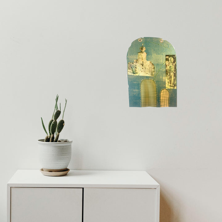 The artwork 'Enter the Temple' features an arch-shaped mixed media collage with turquoise and gold tones, depicting abstract architectural elements and vertical striped patterns. A minimalist white interior setting shows a potted cactus on a modern cabinet beneath the wall art. Created using metallic paper and mixed media techniques by Sunny Chapman.