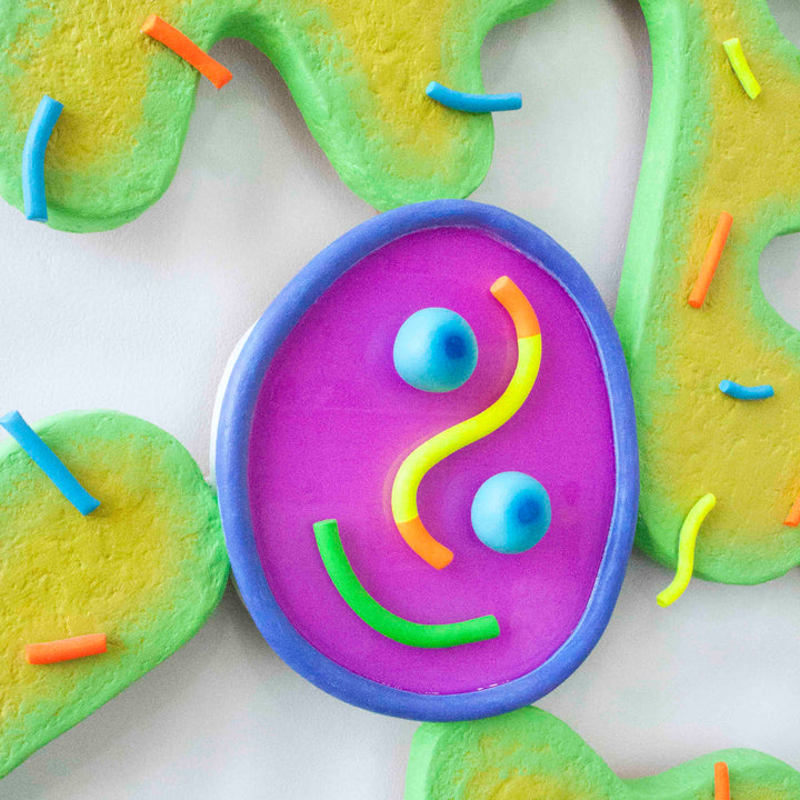 The artwork 'Good Germs' by Ryan Patrick Martin features a playful oval design with a vibrant purple resin pool framed by a blue border. Curved lines in yellow, green, and orange create a whimsical smiling face, surrounded by flowing yellow-green sculptural forms. Multi-colored cylindrical elements accent the composition. Created with wood, epoxy, and acrylic paint.