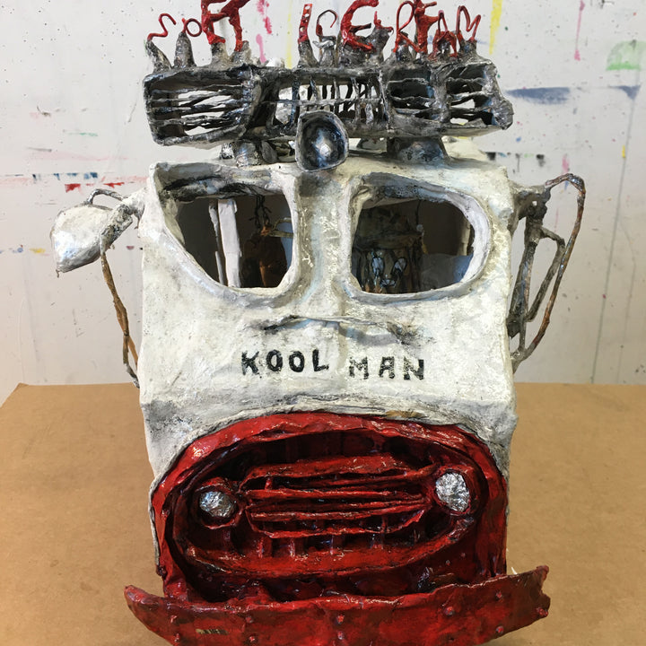 The artwork 'Killing me Softie' by Andrew Chan features a whimsical paper mache sculpture of a character labeled 'Kool Man' with distinctive oversized sunglasses. The piece combines black, white, and red colors, with a textured surface. The sculpture shows a stylized head with a grill-like red mouth and playful text atop, showcasing a raw, street art-inspired aesthetic. - By Andrew Chan