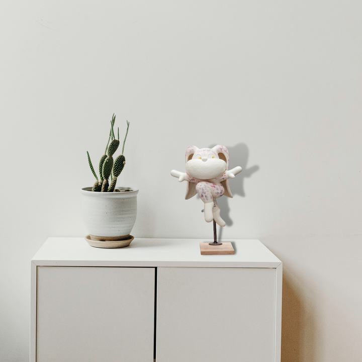 The artwork 'Leap of Faith' by Yusuke Ochiai features a minimalist setting with a white cabinet displaying two items: a small cactus plant in a white ceramic pot with saucer, and a whimsical white cat figurine with wings wearing a pink accessory, mounted on a stand, creating a serene contemporary aesthetic with natural and playful elements.