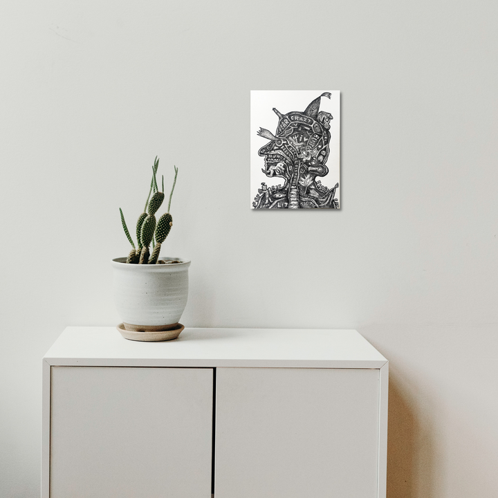 The artwork 'Lit Phrenologist' by Andrew Chan features a black and white ink illustration mounted on a white wall above a minimalist cabinet with a potted cactus. The detailed drawing showcases intricate linework depicting a surreal anatomical study in a phrenological style, rendered in dramatic monochromatic tones. The 12x9 inch watercolor and ink piece contrasts beautifully with the clean modern interior setting.