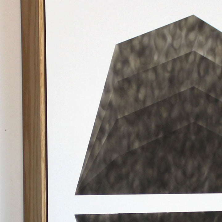 The artwork 'Mild Climate Needed' by Evan Ishmael features a striking geometric composition created with soot on panel. The piece showcases a bold angular form with layered, stepped edges rendered in deep charcoal tones against a stark white background. The soot application creates a textured, atmospheric effect with subtle gradients and shadowy patterns across the geometric surface.